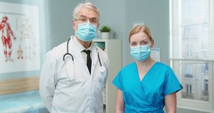Portrait of good-looking medical professionals in protective masks in hospital. Young female nurse and senior male doctor looking at camera in on-call room. Healthcare, pandemic concept.