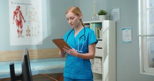 Pretty Caucasian woman in blue medical uniform using tablet in hospital office. Young female nurse swiping, scrolling feed and lbrowsing on Internet. Medicine, technology, digitalization concept.