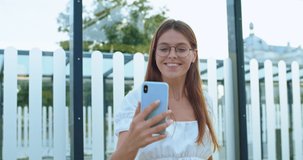 Close up of young woman with long hair in glasses recording video vlog on smartphone camera and smiling cheerfully at street. Portrait of beautiful happy stylish girl influencer vlogger at bus stop.