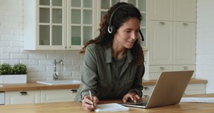 Focused smiling young caucasian woman wearing headphones with microphone, checking home task or discussing educational material with teacher at online video call meeting on computer in kitchen.