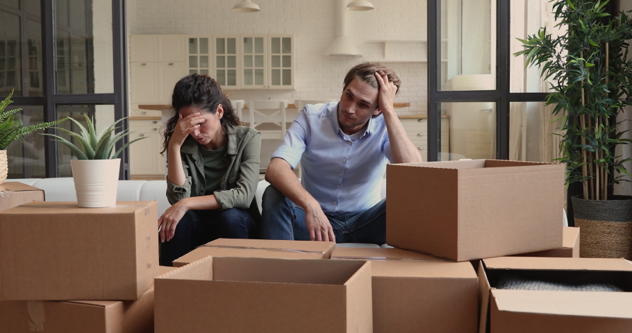 Unhappy young family couple feeling stressed moving into new apartment or moving out of house, sitting together on sofa among huge carton boxes, tired of long renovation relocation processes. Royalty-Free Stock Footage #1066095976