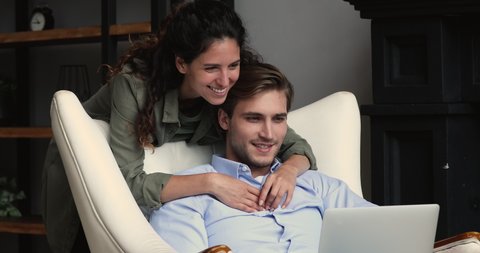 Young man sitting with computer on laps in armchair with smiling pretty wife standing behind, happy family couple involved in online shopping, discussing internet purchases or planning vacation.