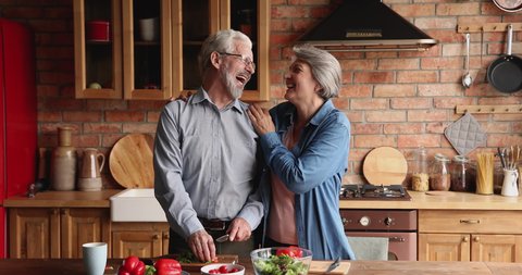 Old spouses enjoy vegetable salad preparation together in kitchen. Caring grey-haired husband feeding loving wife red paprika talking hugging relish romantic date, happy marriage, healthy eat concept Video Stok