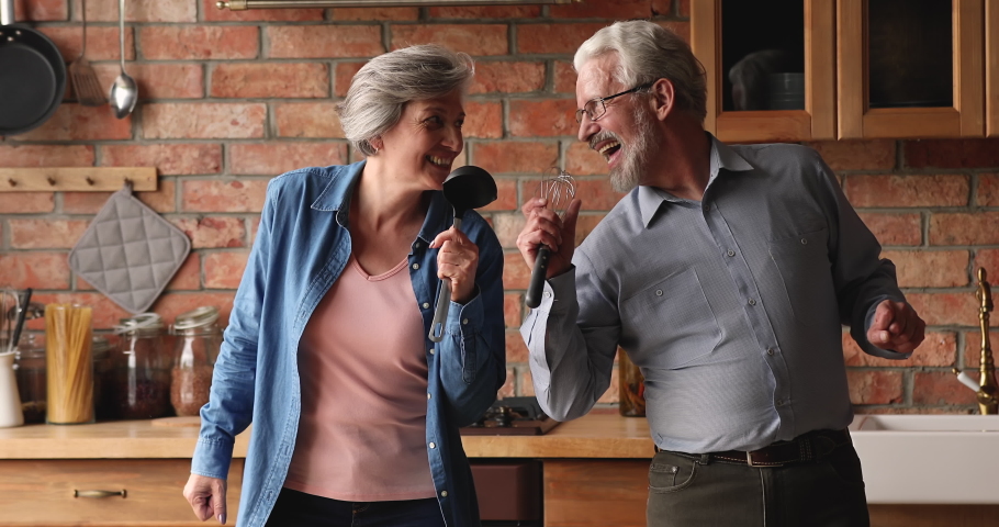 Overjoyed older spouses dancing singing favourite song using ladle and whisk like microphones having fun together in modern kitchen enjoy carefree retired life. Pastime, hobby, karaoke at home concept Royalty-Free Stock Footage #1066100173