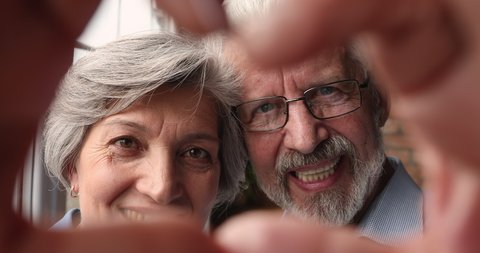 Close up portrait cheerful healthy elderly married couple faces looking through joined fingers making heart shape symbol. Romantic relationships, eternal love, unity, happy long-life marriage concept
