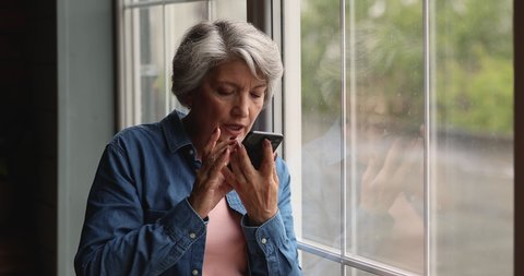 Mature grey-haired woman standing indoor hold smartphone leave voice message using speakerphone and mobile app. Search on internet use voice recognition, speaks commands, ai modern tech usage concept
