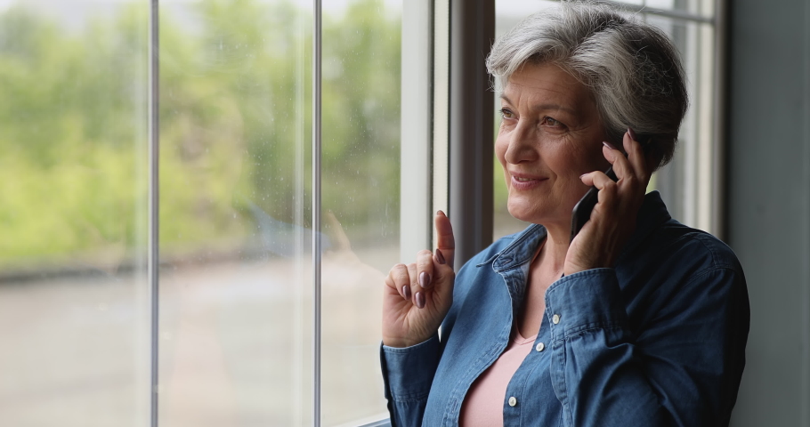 Middle-aged grey-haired woman standing near panoramic window at home holds mobile phone talking. Older generation and modern tech usage, phonecall activity, make order, communication distantly concept Royalty-Free Stock Footage #1066100293