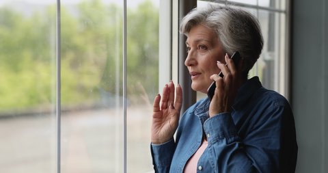 Middle-aged grey-haired woman standing near panoramic window at home holds mobile phone talking. Older generation and modern tech usage, phonecall activity, make order, communication distantly concept