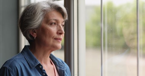Close up view middle-aged 55s grey-haired thoughtful female standing alone indoor looking out the window. Attractive woman with short hairstyle smiling staring at camera having attractive appearance