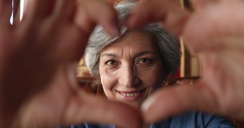 Close up portrait happy smiling mature grey haired woman sit indoor join fingers make heart symbol expressing love show to camera sign of affection. Charity, sincere feelings, video call event concept