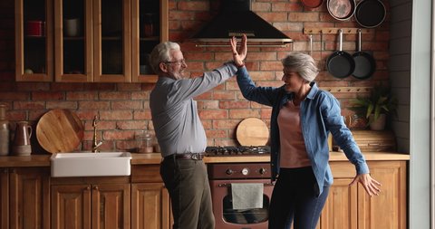 Elderly wife dancing spinning hold hands with husband couple listen music having fun together in cozy kitchen. Romantic date, relations of older generation people. Carefree, active old spouses concept
