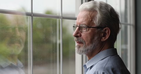 Close up hoary older 65s thoughtful man in glasses standing indoor looking out window. Concept of self-isolation due covid pandemic outbreak, nostalgic mood, yearns, longing for long-gone life moments