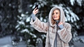 Fashionable lady posing taking selfie in hood fur coat use mobile outdoor at winter forest landscape. Smiling blonde feminine photographing in luxury clothes at snowy trees. Medium shot on RED camera