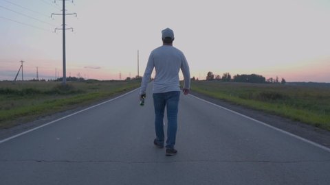 A Man Walking Along The Automobile Road At Sunset, Back View. Young Man in Jeans Walks and Drinks Beer. The concept of Freedom, Happiness, Travel and Free Movement around the World.