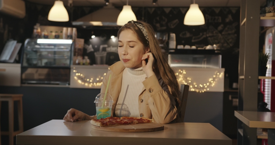 A beautiful young blonde sits in a cafe at a table with slices of pizza on a wooden board and drinks a blue cocktail with lemon through a straw