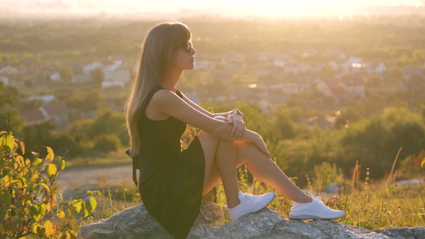 Young pretty woman in black short summer dress sitting on a rock relaxing outdoors at sunset. Fashionable female enjoying warm evening in nature.
