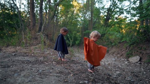 Two little toddler boys cosplay gnomes or hobbits in long capes walking barefoot in green forest. Halloween, kids concept. Amazing fairy tale character. Slow motion. 
