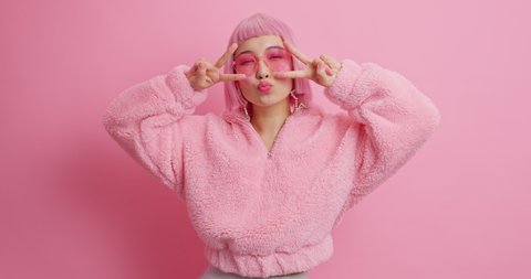 Happy Asian woman makes peace gesture smiles positively has bob hairstyle dressed in winter coat isolated over pink background. Pretty rosy haired female shapes disco hand sign. Body language