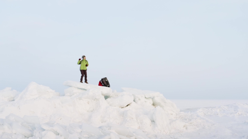 Zooming out view of a happy woman dancing and waving the red scarf on big icy blocks against the blue sky. Seaside. Frozen sea is on the background. Winter