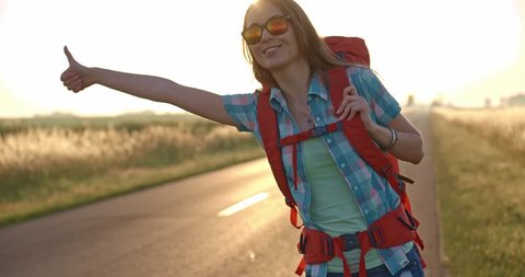 Traveler woman hitchhiking on a sunny road and walking. Young happy backpacker woman looking for a ride to start a journey on a sunlit country road. Slow motion, 120fps. 4K, DCi. Poland, Lower Silesia