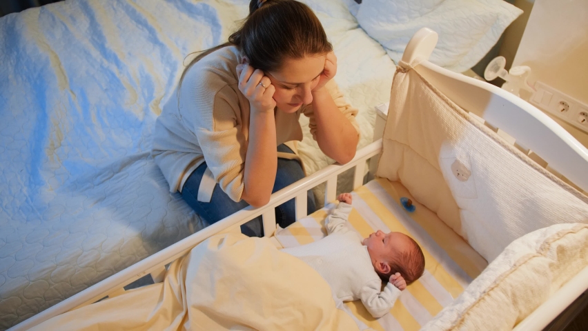 Young tired mother suffering from depression lookin on her sleepless newborn baby in crib at night. Maternal depression after childbirth and sleepless nights