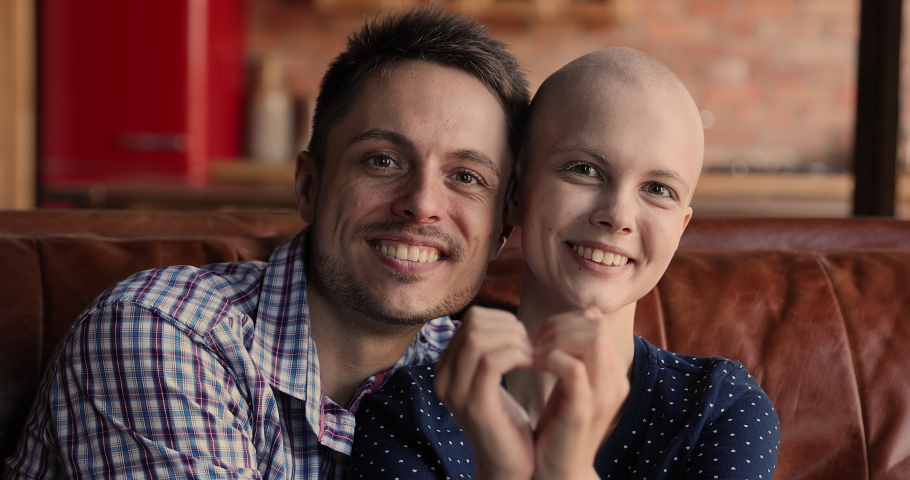 Head shot close up portrait couple husband and bald wife sit on couch smile look at camera, showing with hands heart shape, symbol of encouragement, support to oncology disease cancer patient concept | Shutterstock HD Video #1066120984