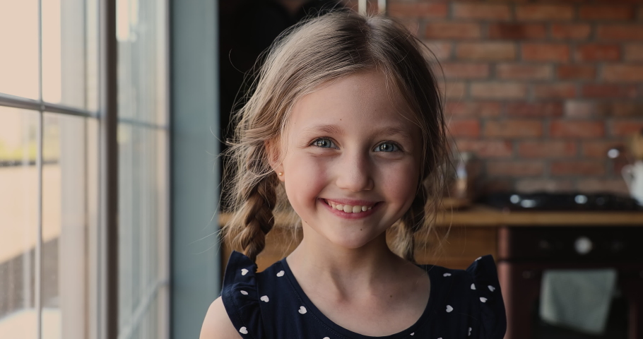 Head shot cute small 7s girl standing in kitchen smiles looks at camera. Adorable caucasian preschool child with pigtail poses indoors. Happy childhood, new Alpha generation offspring portrait concept