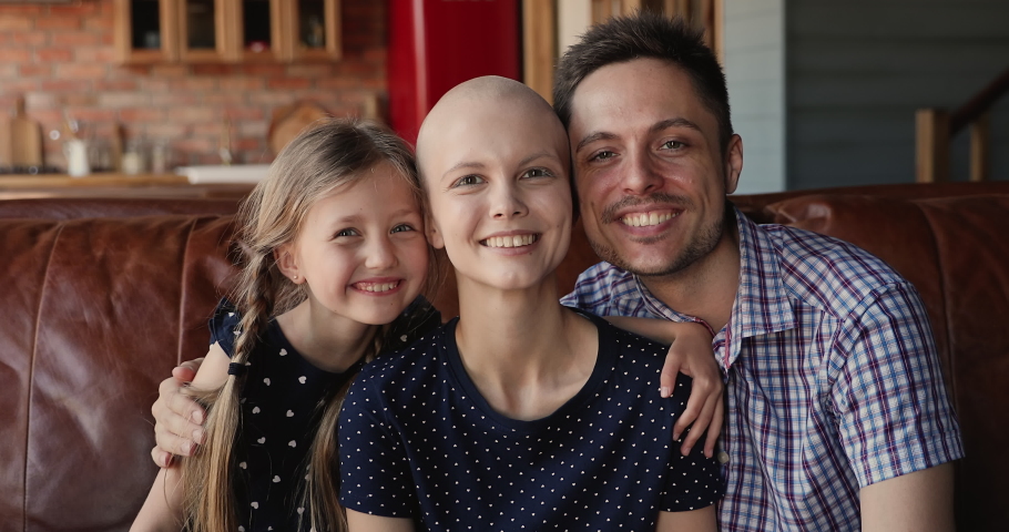 Loving husband, cheerful little daughter hugging young bald female mother wife cancer patient. Happy family celebrate recovery, victory over oncology disease sitting on couch at home feels overjoyed Royalty-Free Stock Footage #1066121122