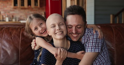 Loving husband, cheerful little daughter hugging young bald female mother wife cancer patient. Happy family celebrate recovery, victory over oncology disease sitting on couch at home feels overjoyed