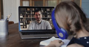 On-line male tutor teaches little girl by videocall due covid-19, home-schooling concept. View over kid shoulder sit at table wear headphones listens teacher explanation writes down exercise solution