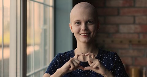 Head shot portrait young 20s bald woman standing indoor smile look at camera showing with fingers heart shape. Symbol of gratitude for full oncology disease recovery, supporting cancer patient concept