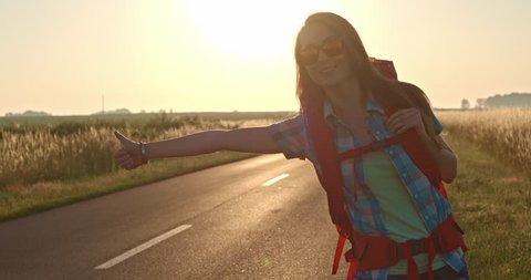 Traveler woman hitchhiking on a sunny road and walking. Young happy backpacker woman looking for a ride to start a journey on a sunlit country road. Slow motion, 120fps. 4K, DCi. Poland, Lower Silesia
