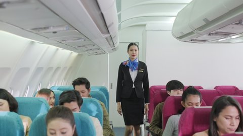 Portrait of attractive stewardess in uniform walking in the economy class of airplane and smile. Passengers sitting and fasten seat belt for safety flight. Transportation and service concept.