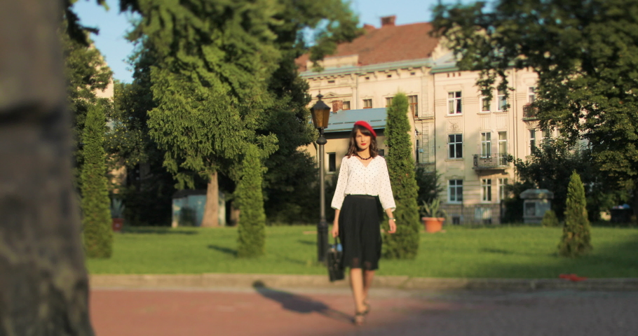 Pretty Caucasian happy young stylish woman in red beret walking outdoor in city garden. Beautiful cheerful girl on a walk alone in good mood. French style. Paris fashion. Outdoors concept