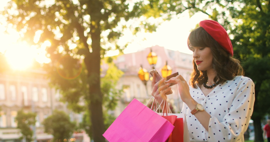 Portrait of young Caucasian pretty girl with shopping bags buying on internet on smartphone while standing on street. Beautiful female paying with credit card on mobile phone with funny shocked face