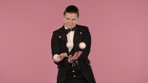 Young man in a black and white suit juggles with three balls. Circus performer practices concentration, skill and reaction in the studio on pink background. Isolated close up. Slow motion.