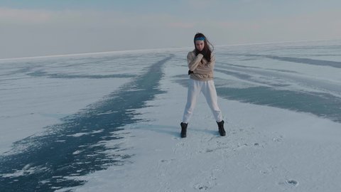 Stylish young woman, professional dancer, dancing energetic jazz funk or hip hop on frozen lake in cold winter. Female hipster dances challenge for social networks on snow-covered frozen arctic river