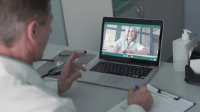 Old male doctor talking to senior female patient by tele medicine online video call in clinic. Physician using telehealth medical chat virtual healthcare visit on laptop screen. Over shoulder view