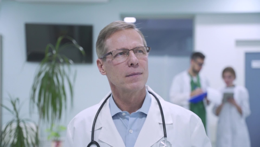 Confident old medical professional doctor walking in hospital hallway with medic staff greeting nurse. Senior male physician wears white coat, stethoscope and glasses going along clinic corridor. Royalty-Free Stock Footage #1066143373