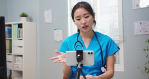 Charming female nurse making video blog about health protection during pandemic. Young Asian woman talking to viewers, followers, live streaming from hospital. Medicine, social media concept.