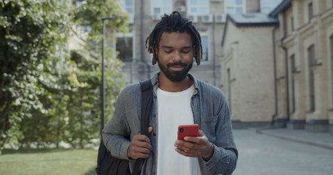 Bearded young man with dreadlocks using smartphone and smiling. Cheerful guy in casual clothes carrying bag on his shoulder and chatting on social media while walking on city street.