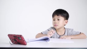 Asian boy about 5 years old writing while watching a mobile phone as studying online due to Covid 19 pandemic