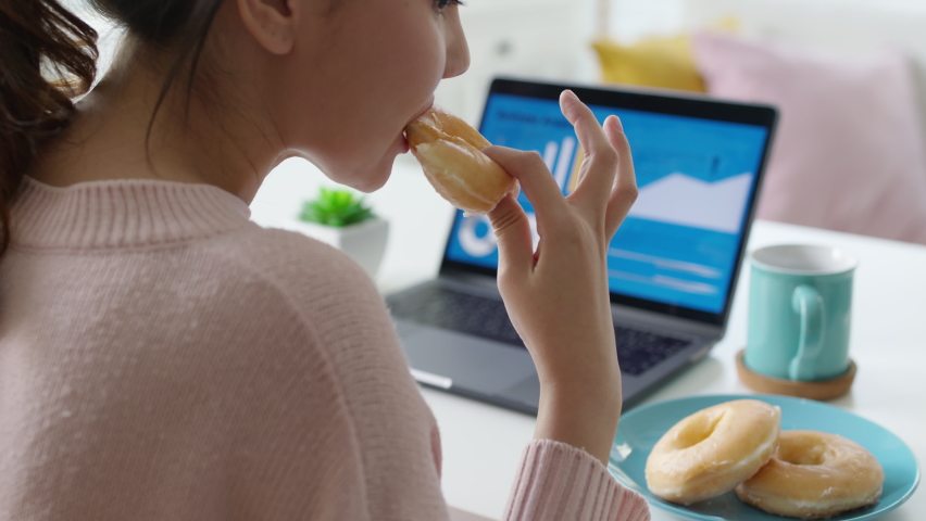 Young attractive beautiful asian female hungry eat doughnut take away snack food with full mouth look at computer notebook at home in busy work from home multitask unhealthy meal lifestyle concept. | Shutterstock HD Video #1066146721