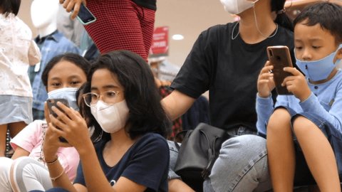 Asian children wearing face masks sitting on the stairs staring into their smartphones in shopping mall. Bali-December-2020