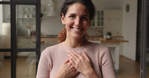 Smiling sincere kind young caucasian woman holding folded hands on heart chest, feeling deeply thankful at home. Happy inspired beautiful millennial lady showing gratitude, expressing appreciation.