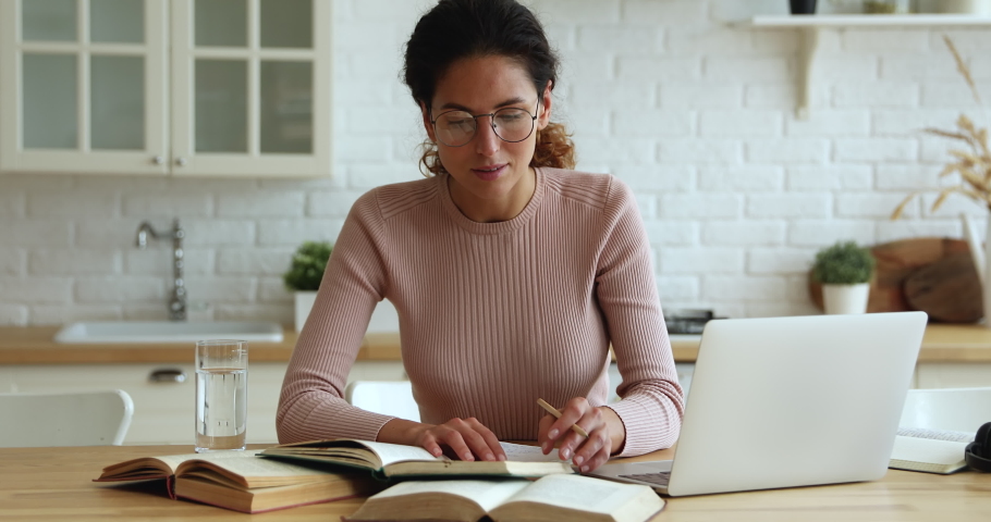 Concentrated young smart woman in eyeglasses sitting at table with books and computer, searching and highlighting important information in educational literature, preparing for exams in kitchen. Royalty-Free Stock Footage #1066149838