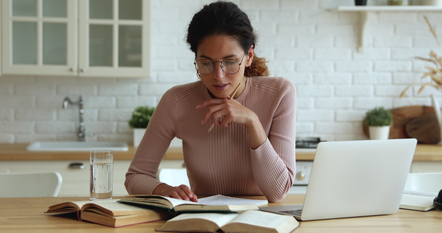Concentrated young smart woman in eyeglasses sitting at table with books and computer, searching and highlighting important information in educational literature, preparing for exams in kitchen. Royalty-Free Stock Footage #1066149838