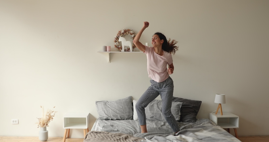 Full length overjoyed millennial caucasian fit woman dancing to favorite disco music, jumping on bed, enjoying free weekend leisure time alone in bedroom, feeling cheerful while celebrating freedom. Royalty-Free Stock Footage #1066150153