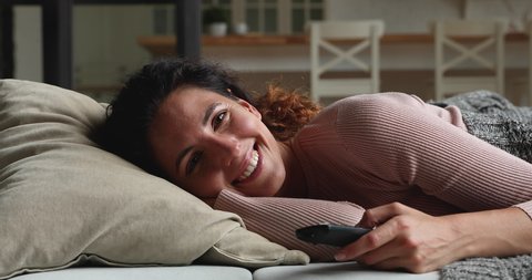 Happy young beautiful woman lying on comfortable sofa under warm blanket, watching tv shows or laughing at funny comedian movie using remote controller at home, enjoying spending weekend lazy time.