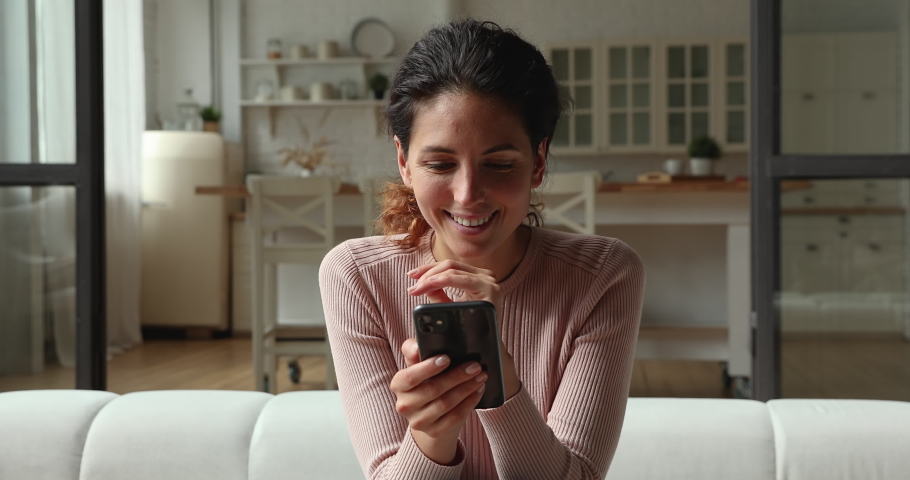 Head shot happy millennial pretty woman sitting on comfortable sofa, involved in chatting in dating application or communicating distantly with friends using smartphone at home, leisure hobby concept. | Shutterstock HD Video #1066150192
